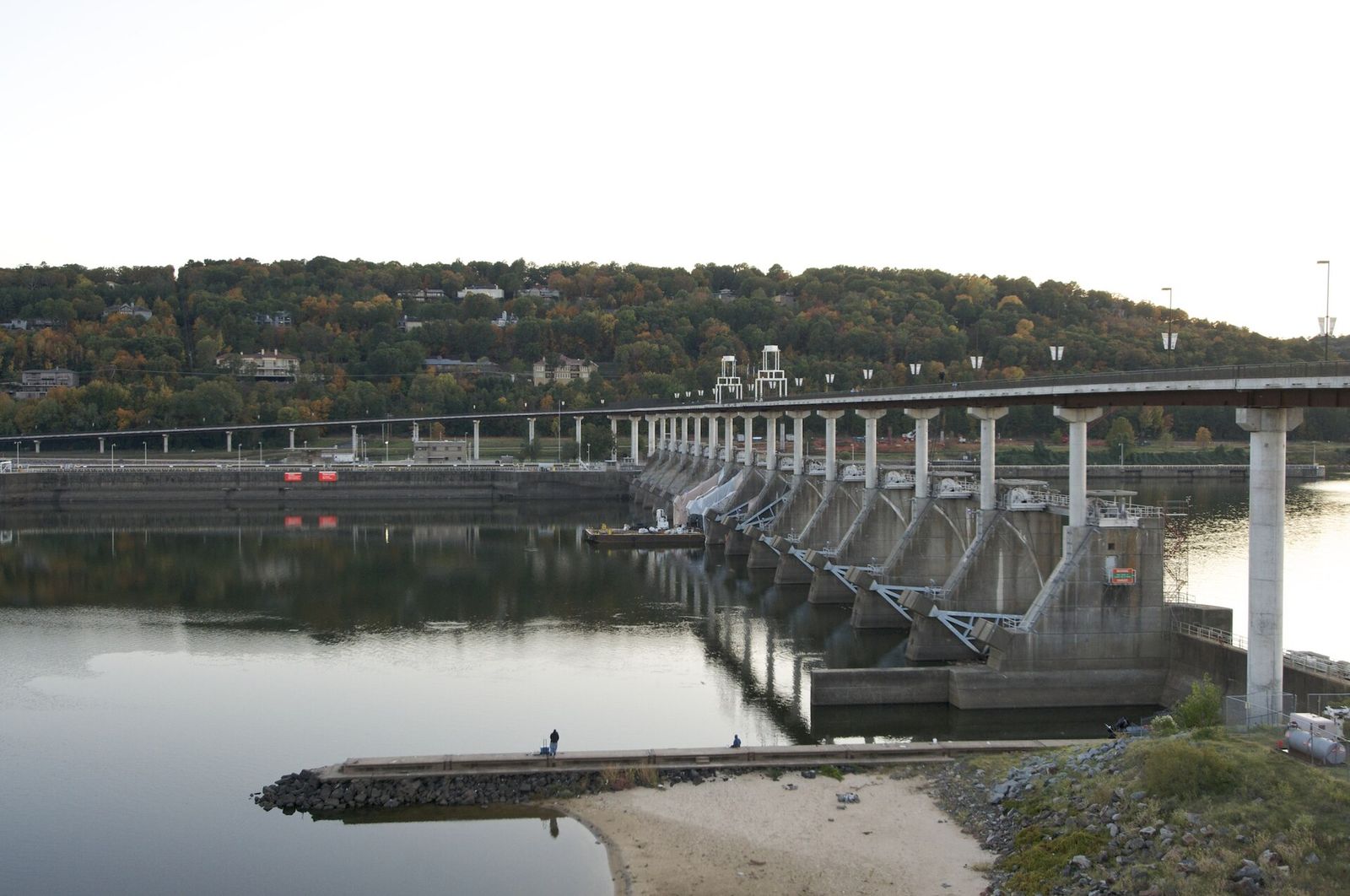 Anglers fishing from the bank below a dam at dusk, with the spillway gates and a bridge behind them.